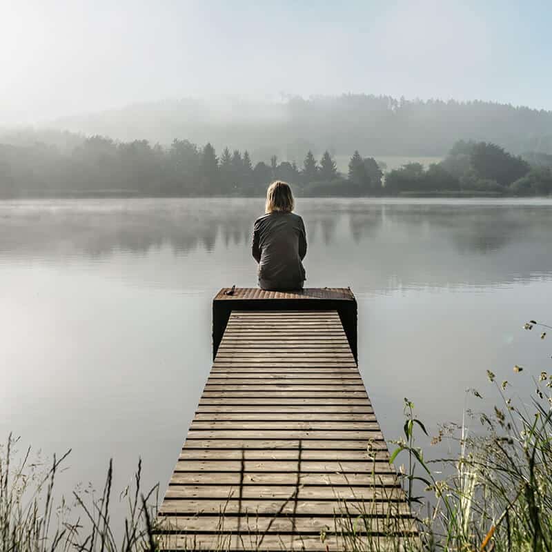 Woman sitting on a wooden dock overlooking a lake on a foggy day.